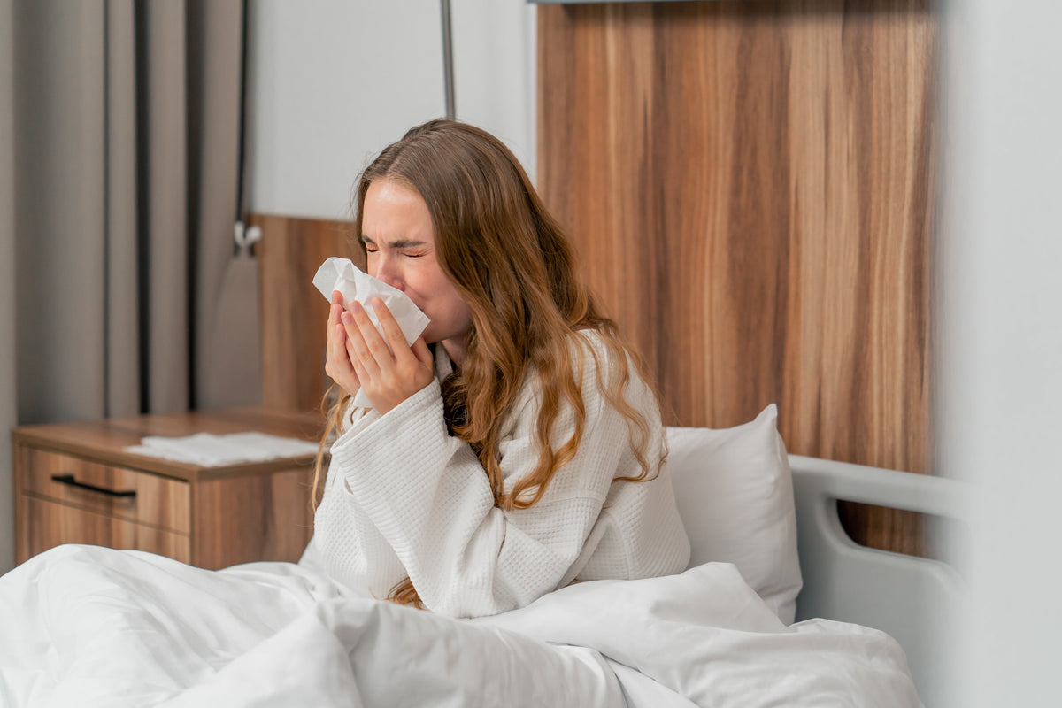 Vrouw in badjas niest in een tissue terwijl ze in bed zit, mogelijk door huisstofmijtallergie.